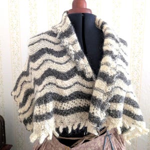 May include: A hand-knitted shawl in a chevron pattern, displayed on a mannequin. The shawl features alternating bands of grey and cream-coloured yarn. The edges are finished with a scalloped trim. The shawl is a cosy accessory.
