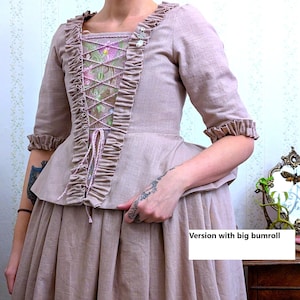 May include: A dusty rose linen dress with a fitted bodice and a full skirt. The bodice features a lace-up front with a floral embroidery design and a ruffle trim. The dress has a gathered skirt and a large bumroll.
