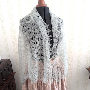 May include: A delicate, light gray shawl with an intricate lace pattern is draped over a mannequin. The shawl has a scalloped edge and a flowing, airy design. The mannequin is on a pink dress, and the background is a floral wallpaper.