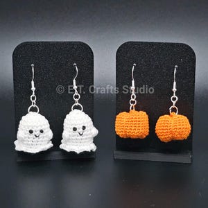 May include: Two pairs of dangle earrings featuring crocheted charms. One pair consists of white ghosts with black embroidered eyes and mouths. The other pair features orange pumpkins with textured surfaces. All charms hang from silver-toned metal hooks.