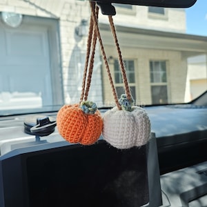 May include: Two crocheted pumpkin ornaments hanging from a rearview mirror. One pumpkin is bright orange and the other is off-white. Both feature a small, textured green stem at the top and are suspended by thin, brown crocheted yarn strings.