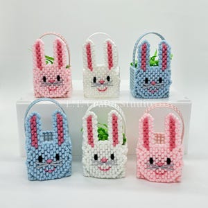 May include: Six small plastic Easter baskets with bunny faces. The baskets are in pink, blue, and white. Each basket has a handle and is filled with green grass.