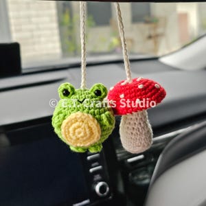 May include: A green crocheted frog holding a yellow crocheted flower and a red and white crocheted mushroom hanging from a rearview mirror in a car.