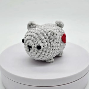 May include: A gray crocheted plush toy shaped like a bear with black eyes and a red felt heart on its side.