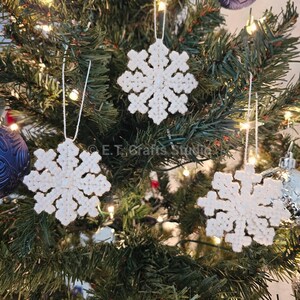 May include: Three white snowflake ornaments hanging on a Christmas tree. The ornaments are made of a textured material and have a detailed snowflake design. The tree is decorated with other ornaments and lights. The text "E.T. Crafts Studio" is visible.