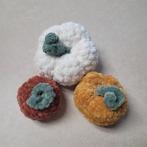 May include: Three crocheted pumpkins in different colors: white, brown, and orange. Each pumpkin has a green crocheted stem.