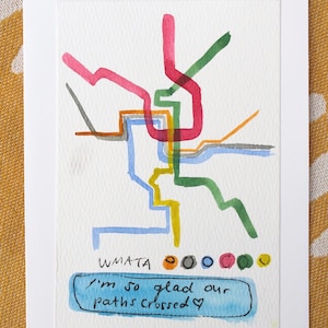 May include: A white greeting card featuring a watercolor illustration of a transit map with intersecting lines in pink, green, blue, yellow, orange, and gray. Below the map, the text WMATA appears with colored dots and the handwritten phrase I'm so glad our paths crossed with a small heart symbol.