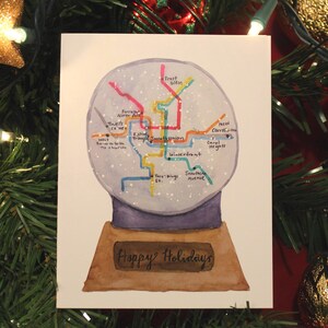 May include: Watercolor Christmas card featuring a snow globe design with a map inside. The map has colorful lines and labels. The base of the card reads "Happy Holidays". The card is surrounded by Christmas tree branches and ornaments.
