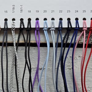 May include: A color chart featuring a horizontal white display card with numbered slots from 8 to 33, holding various thin, flexible cords in a wide spectrum of colors including yellow, gray, black, white, purple, blue, pink, red, and green.