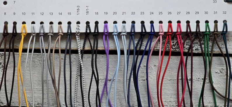 May include: A color chart featuring a horizontal white display card with numbered slots from 8 to 33, holding various thin, flexible cords in a wide spectrum of colors including yellow, gray, black, white, purple, blue, pink, red, and green.
