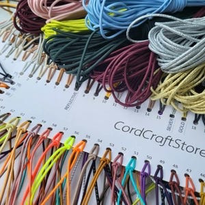 May include: A white display card labeled CordCraftStore featuring a wide variety of colorful cords organized by number. The collection includes numerous shades such as neon yellow, light blue, silver, gold, black, and burgundy, all neatly looped.