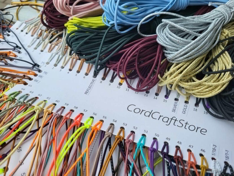 May include: A white display card labeled CordCraftStore featuring a wide variety of colorful cords organized by number. The collection includes numerous shades such as neon yellow, light blue, silver, gold, black, and burgundy, all neatly looped.