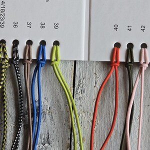 May include: A color sample card featuring a series of numbered holes with attached cords in various colors and patterns, including black, yellow, blue, green, orange, pink, and brown, arranged in a row on a white background with printed numbers.