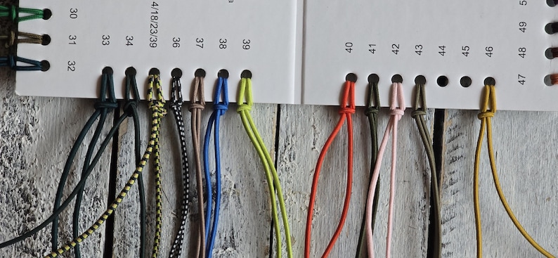 May include: A color sample card featuring a series of numbered holes with attached cords in various colors and patterns, including black, yellow, blue, green, orange, pink, and brown, arranged in a row on a white background with printed numbers.