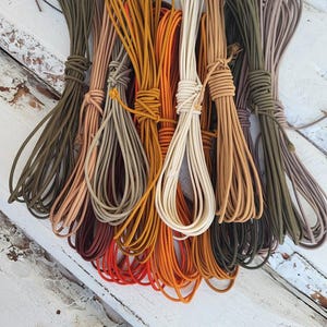 May include: A collection of multiple bundles of smooth, round cord in various earth tones and vibrant colors, including olive green, tan, mustard yellow, bright orange, cream, and brown, all neatly coiled and tied with matching cord segments.