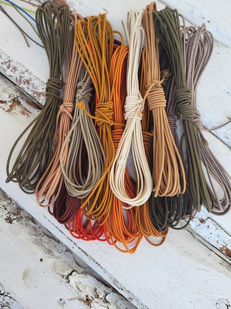 May include: A collection of multiple bundles of smooth, round cord in various earth tones and vibrant colors, including olive green, tan, mustard yellow, bright orange, cream, and brown, all neatly coiled and tied with matching cord segments.