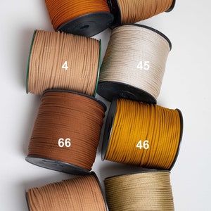 Peut inclure: Huit bobines de cordon dans diverses nuances de brun, beige et doré, chacune marquée d'un numéro blanc : 65, 5, 4, 45, 66, 46, 6 et 44. Les cordons sont étroitement enroulés sur des bobines en plastique noir, montrant une texture tissée.