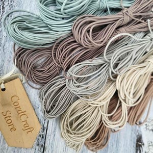 May include: A collection of coiled elastic cords in various neutral shades including sage green, dusty rose, taupe, grey, and cream. A light-colored wooden tag with the laser-engraved text CordCraft Store is attached to the bundles of cord.