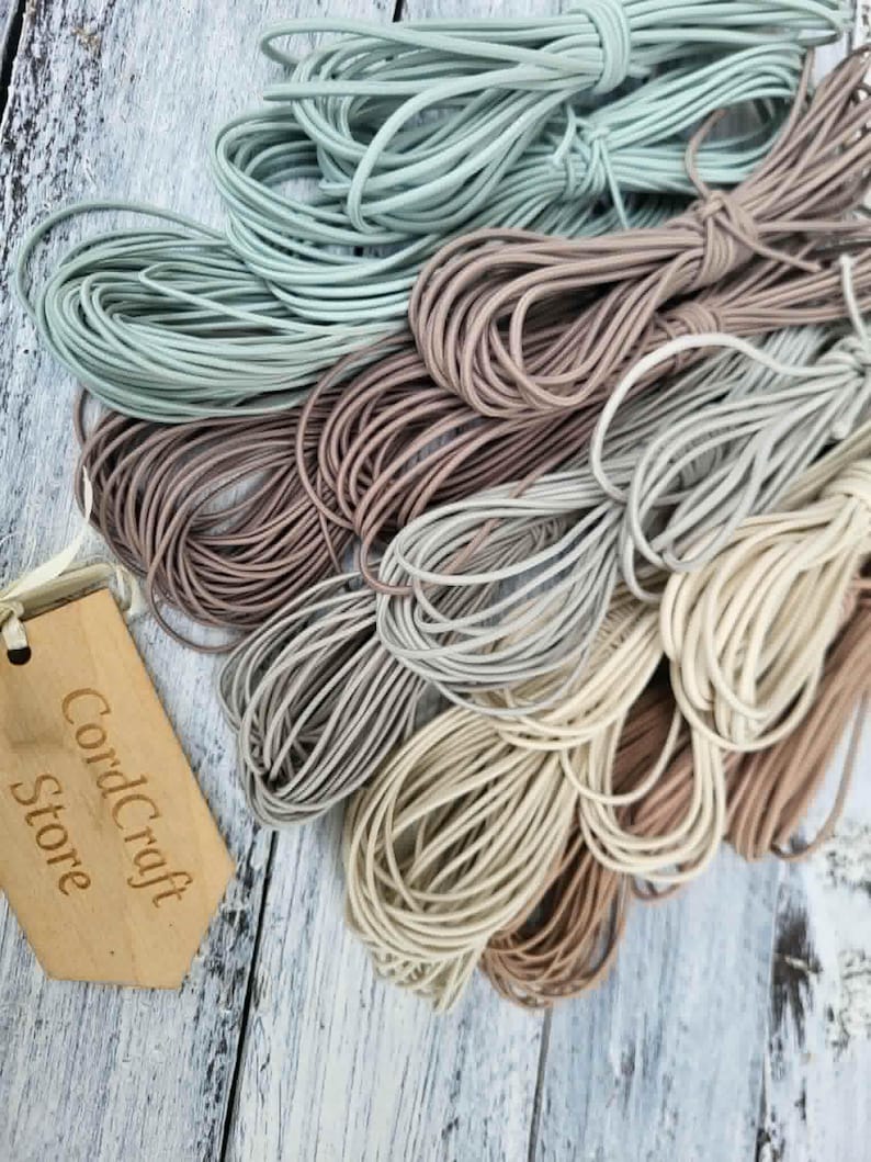 May include: A collection of coiled elastic cords in various neutral shades including sage green, dusty rose, taupe, grey, and cream. A light-colored wooden tag with the laser-engraved text CordCraft Store is attached to the bundles of cord.