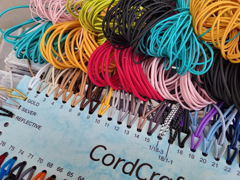 May include: A color sample card labeled CordCraft featuring numerous loops of elastic cord in various shades including black, white, pink, yellow, teal, red, and brown. The card includes numbered slots and labels for gold, silver, and reflective cords.
