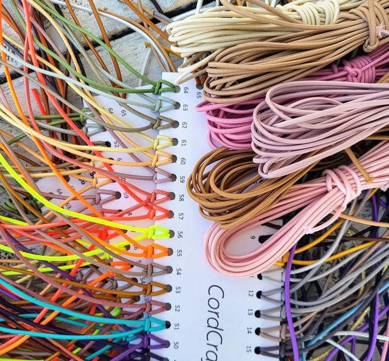 May include: A color sample card labeled CordCraft with numbered slots holding various lengths of thin, flexible elastic cord in a wide spectrum of colors including neon yellow, orange, pink, brown, beige, green, blue, and purple.