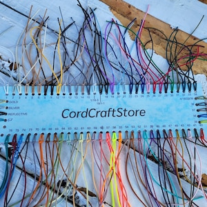 May include: A light blue rectangular display card labeled CordCraftStore featuring numerous numbered holes, each threaded with a different colored cord. The cords vary in shades including white, yellow, pink, red, blue, green, and black.