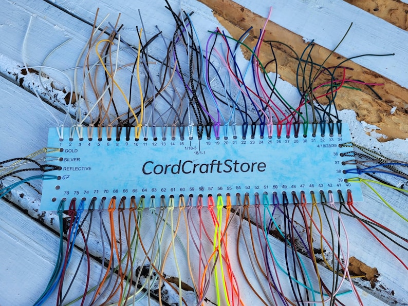 May include: A light blue rectangular display card labeled CordCraftStore featuring numerous numbered holes, each threaded with a different colored cord. The cords vary in shades including white, yellow, pink, red, blue, green, and black.