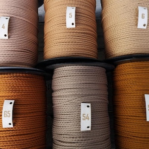 May include: Spools of cord in various shades of brown and tan. Each spool has a small tag with a number on it. The colors range from light beige to a deep rust color. The cord appears to be made of a woven material.