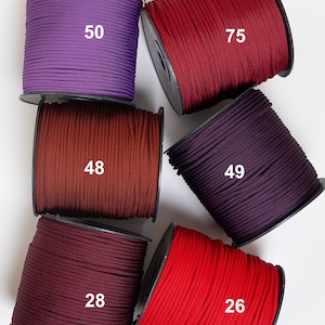 Op de afbeelding: Zes klossen koord in verschillende kleuren, waaronder paars, donkerrood, roestbruin, diep pruim, bordeauxrood en felrood. Elke klos is voorzien van een wit nummer: 50, 75, 48, 49, 28 en 26, wat de specifieke kleurvariant van elk koord aangeeft.