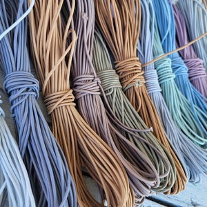 May include: A collection of multiple bundles of thin, round elastic cord arranged side by side. The cords are available in a variety of muted colors including white, light blue, tan, mauve, olive green, brown, teal, and dusty purple.