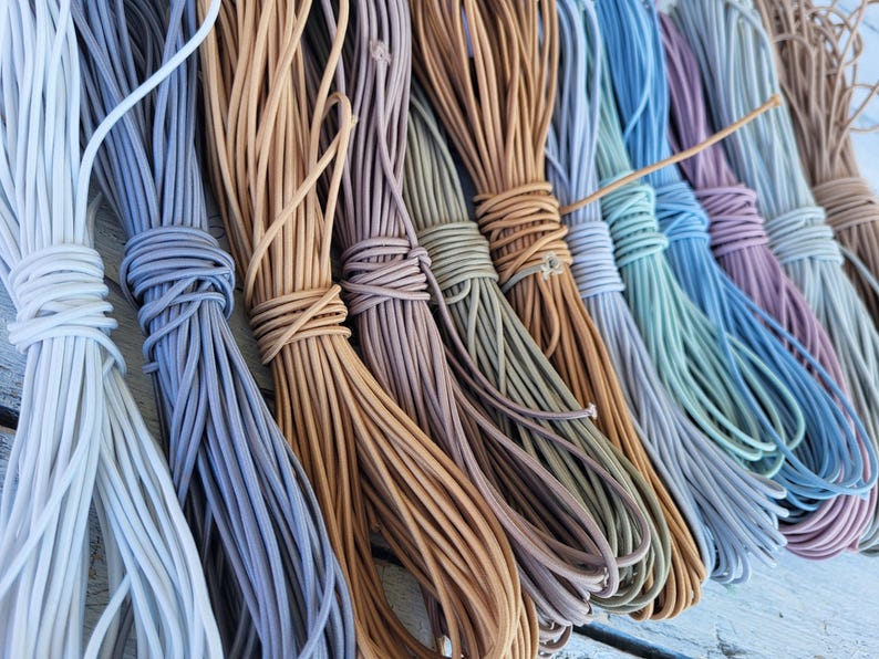 May include: A collection of multiple bundles of thin, round elastic cord arranged side by side. The cords are available in a variety of muted colors including white, light blue, tan, mauve, olive green, brown, teal, and dusty purple.