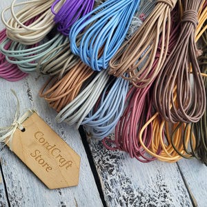 Puede incluir: Una colección de cordones de cuero multicolores agrupados en varios tonos, incluyendo rosa, azul, beige, marrón y verde. Una etiqueta de madera clara con el texto CordCraft Store está unida a los cordones con un cordel fino y claro.