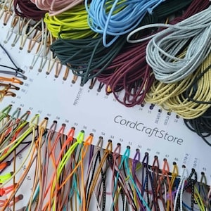 May include: A white display card labeled CordCraftStore featuring a wide variety of colorful elastic cords organized by numbered slots. The cords come in diverse shades including neon yellow, light blue, silver, gold, black, and deep red.