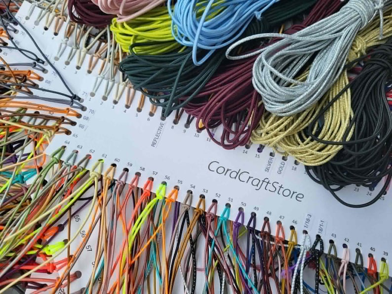 May include: A white display card labeled CordCraftStore featuring a wide variety of colorful elastic cords organized by numbered slots. The cords come in diverse shades including neon yellow, light blue, silver, gold, black, and deep red.