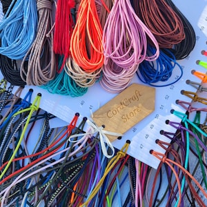 May include: A collection of multi-colored cords organized on a white display card with numbered slots. The cords feature various solid colors, patterns, and textures. A wooden tag with the text CordCraft Store is attached to the center.
