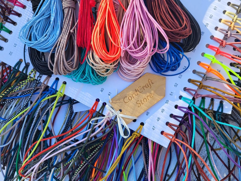May include: A collection of multi-colored cords organized on a white display card with numbered slots. The cords feature various solid colors, patterns, and textures. A wooden tag with the text CordCraft Store is attached to the center.