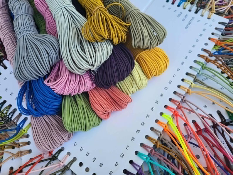 May include: A color chart featuring numerous bundles of thin, elastic cord in a wide variety of colors including grey, blue, pink, green, yellow, and purple. The cords are attached to a white display card with numbered holes for identification.