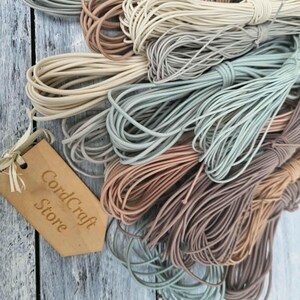 May include: A collection of coiled elastic cords in various muted earth tones including beige, tan, sage green, and dusty rose. A light brown wooden tag with the text CordCraft Store is attached to the cords with a thin piece of string.