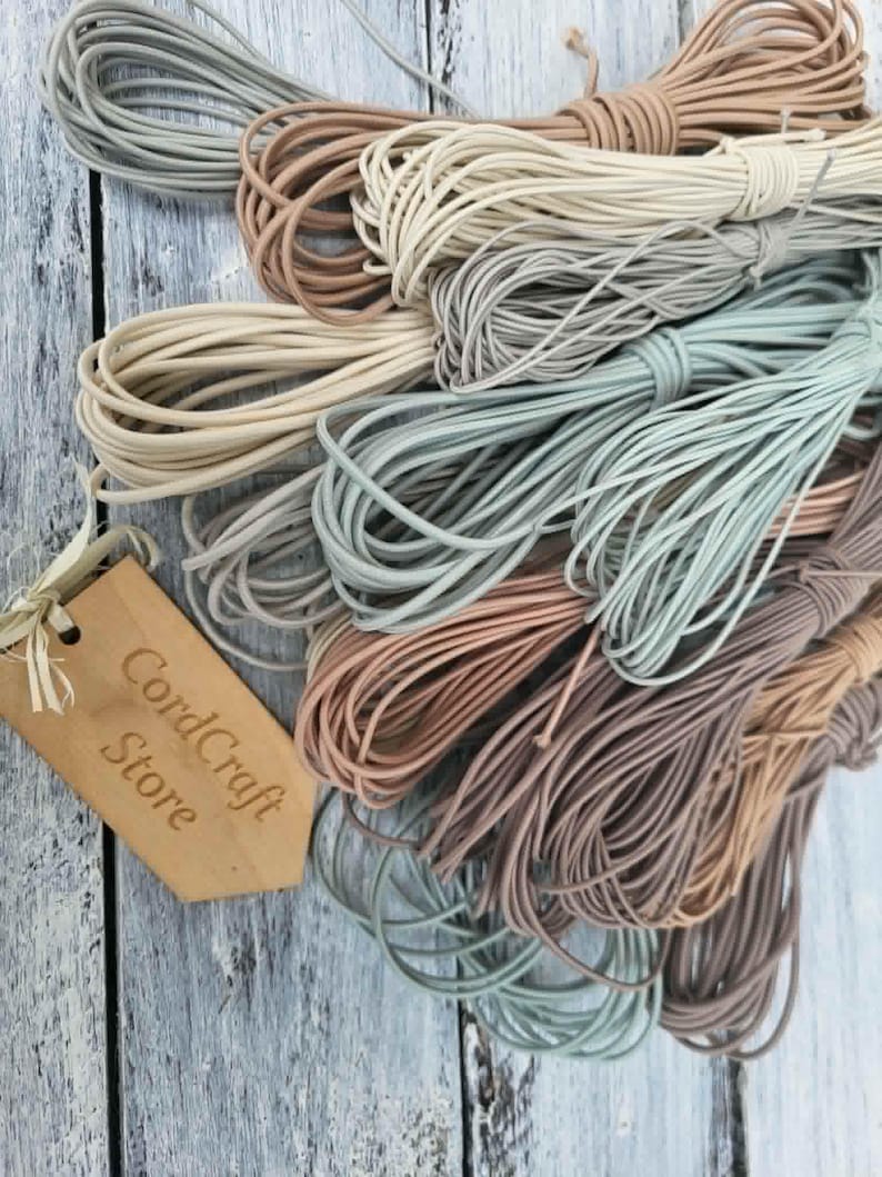 May include: A collection of coiled elastic cords in various muted earth tones including beige, tan, sage green, and dusty rose. A light brown wooden tag with the text CordCraft Store is attached to the cords with a thin piece of string.