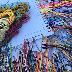 Puede incluir: Una colección de cordones y cuerdas coloridas, incluyendo amarillo, verde, rojo y azul, están dispuestas sobre una tarjeta blanca con agujeros numerados. Los cordones son probablemente para manualidades o la fabricación de joyas.