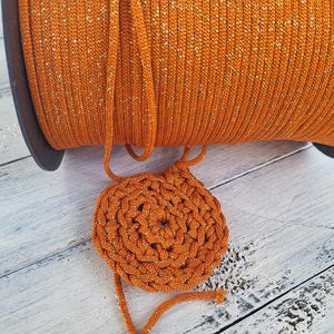May include: A large spool of vibrant orange cord with subtle gold metallic flecks wound tightly around a dark circular core, with a small, circular, hand-crocheted piece made from the same orange and gold cord resting in front of the spool.