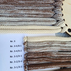 Puede incluir: Una colección de cordones trenzados en varios tonos de marrón, beige y blanco, organizados en una bobina de madera y una tarjeta blanca. La tarjeta presenta etiquetas impresas que incluyen Nr. 3, Nr. 3-5/8-1, Nr. 3-4/8-2, Nr. 3-3/8-3, Nr. 3-2/8-4, Nr. 3-1/8-5 y Nr. 8.