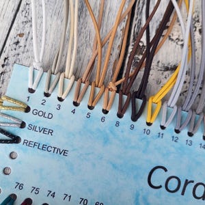 Op de afbeelding: Een lichtblauwe displaykaart met het label CordCraft met een verscheidenheid aan elastische koorden in kleuren zoals wit, beige, bruin, geel, grijs en zwart. De kaart bevat genummerde sleuven van 1 tot 15 en labels voor goud, zilver en reflecterend.