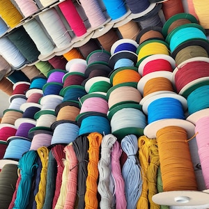 May include: A collection of numerous spools of elastic cord in a wide variety of solid colors, including shades of blue, red, yellow, green, pink, orange, black, and white. The spools are arranged in rows, with some loose bundles of cord in the foreground.