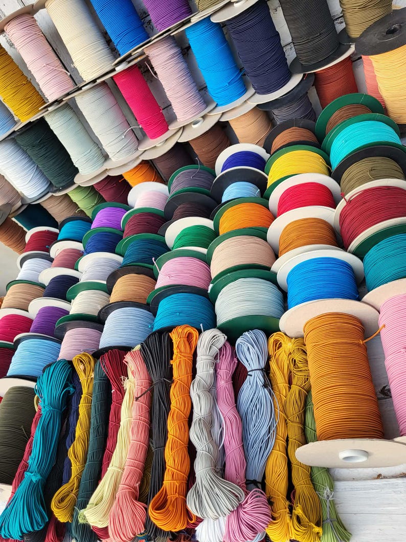 May include: A collection of numerous spools of elastic cord in a wide variety of solid colors, including shades of blue, red, yellow, green, pink, orange, black, and white. The spools are arranged in rows, with some loose bundles of cord in the foreground.