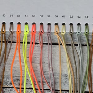 May include: A white color chart card featuring a series of numbered holes from 46 to 73, each holding a looped cord in various colors including brown, purple, teal, orange, neon yellow, pink, beige, grey, green, and black, labeled as REFLECTIVE.