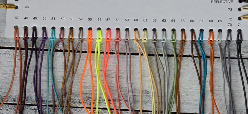 May include: A white color chart card featuring a series of numbered holes from 46 to 73, each holding a looped cord in various colors including brown, purple, teal, orange, neon yellow, pink, beige, grey, green, and black, labeled as REFLECTIVE.