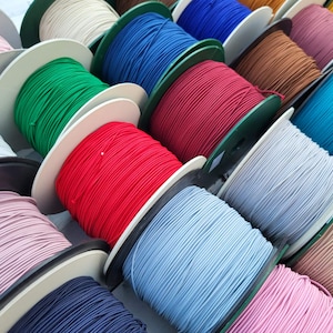 May include: A collection of multiple spools of elastic cord in a variety of solid colors including red, blue, green, pink, yellow, purple, brown, gray, and black. Each spool features a circular plastic frame holding the tightly wound cord.