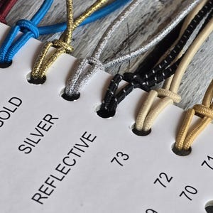 May include: A white display card featuring numbered holes and labels including 74, GOLD, SILVER, and REFLECTIVE. Various elastic cords in colors such as maroon, blue, gold, silver, black with white flecks, and beige are looped through the holes.