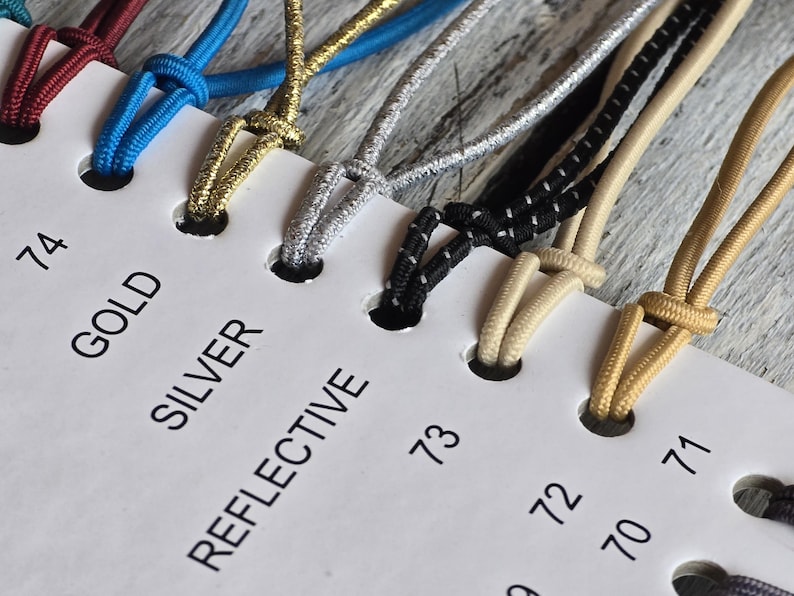 May include: A white display card featuring numbered holes and labels including 74, GOLD, SILVER, and REFLECTIVE. Various elastic cords in colors such as maroon, blue, gold, silver, black with white flecks, and beige are looped through the holes.